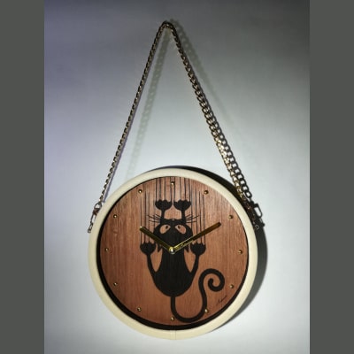 Orologio da parete in legno di noce con incisione a laser di un gatto che graffia la parete. Dettagli dorati su lancette e numeri, cornice in ecopelle beige e tracolla in metallo dorato. Articolo artigianale, unico e raffinato, perfetto come regalo originale per ogni occasione.