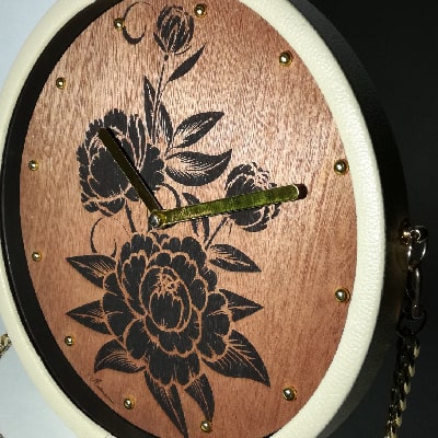 Particolare: Orologio moderno con incisione di romantiche rose su noce, lancette dorate, cornice in ecopelle beige e tracolla dorata. Eleganza artigianale per ogni spazio.